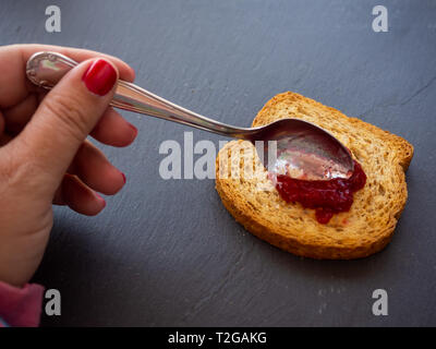 Une femme avec des ongles peints en rouge de smearing Confiture de framboises avec une cuillère sur une tranche de pain grillé Banque D'Images