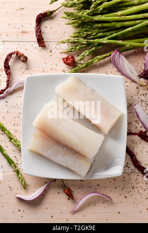 Portrait de quelques tranches de matières de la morue sur une plaque en céramique blanc, quelques asperges sauvages, quelques piments et quelques tranches d'oignon violet, sur un Banque D'Images