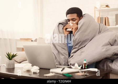 Sick black student le travail à domicile sur ordinateur portable avec le devoir ou projet universitaire, enroulée sous couverture sur la table, l'espace libre Banque D'Images
