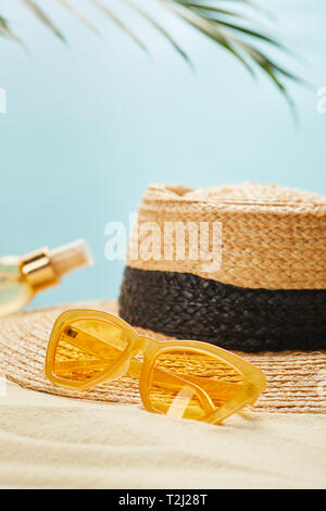 Focus sélectif de lunettes de près de chapeau de paille et une bouteille avec de l'huile de bronzage sur la plage de sable isolée sur bleu Banque D'Images