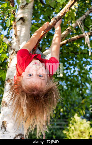 Fille d'âge élémentaire accrochée à une branche d'arbre tout en jouant dans un jardin d'été - la sécurité des enfants à risque ou concept de jeu Banque D'Images