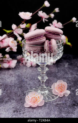 Macaron français dessert cookies rose lilas dans un vase verrerie sur fond noir avec des fleurs roses Banque D'Images