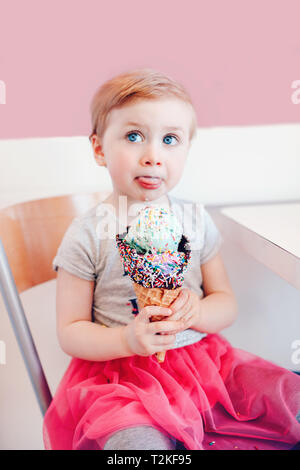 Funny Cute blonde caucasienne babyl petite fille aux yeux bleus, de lécher la crème glacée manger dans un grand cornet gaufré avec sprinkles et montrant la langue. Heureux chi Banque D'Images