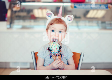 Adorable mignon drôle Caucasian blonde enfant aux yeux bleus portant bandeau unicorn manger lécher la crème glacée dans un grand cornet gaufré avec paillettes. Banque D'Images