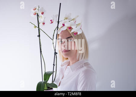 Jeune femme tenant une fleur d'orchidée dans un pot isolé sur fond blanc en studio. Girl looking at camera. Close up, selective focus Banque D'Images