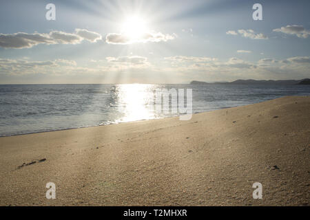 La plage d'or le long de la mer bleu calme sous un ciel clair avec des nuages blancs et le soleil brillant Banque D'Images