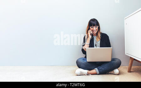 Businesswoman sitting on floor looking at a laptop computer de conversations au téléphone mobile. Femme entrepreneur travaillant à domicile on cell phone usin Banque D'Images