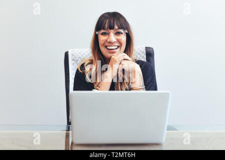 Close up of a smiling woman assise à son bureau en bureau avec un ordinateur portable à l'avant. Cheerful businesswoman in office working on laptop. Banque D'Images