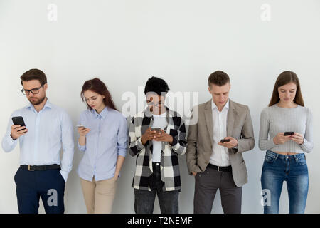 Groupe d'étudiants ou businesspeople standing dans une rangée en face de mur blanc, tenant à la recherche sur les smartphones, en attente d'entrevue. Chat dans la n Banque D'Images