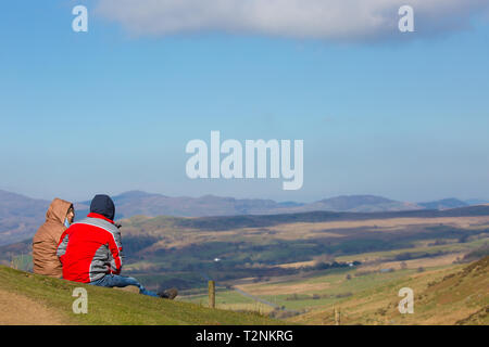 Vue arrière du couple sur une montagne en Mid-Wales siégeant ensemble on grassy hillside in sunshine, regardant la vue paysage spectaculaire en face d'eux. Banque D'Images