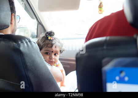 Une belle fille bébé indien enfant assis dans un siège avant de voiture est à la recherche de quelque chose sur le siège arrière d'une voiture en mouvement. Le mode de vie des Indiens Banque D'Images