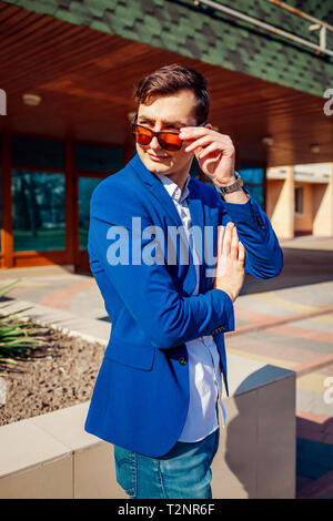 Heureux jeune homme élégant en ville à pied. Beau gars portant des vêtements et accessoires classique et enlever les lunettes. Spring street fashion. Banque D'Images