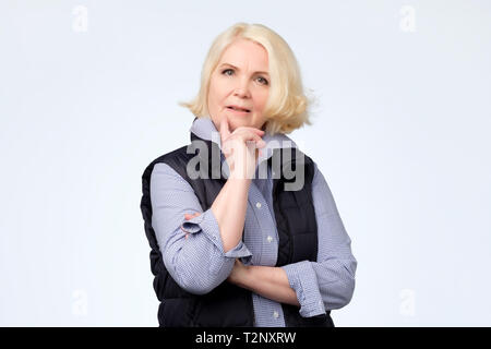 Troublé et concerné senior woman sourire essayant de penser créer un plan dans l'esprit sur la mur gris. Studio shot Banque D'Images