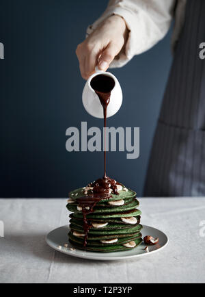 Woman pouring chocolat chaud sur les crêpes maison verte avec les épinards, la banane et l'avoine sur fond gris au petit déjeuner Banque D'Images