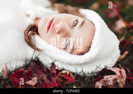 Fille en haut à capuchon blanc couché sur l'herbe entre les feuilles d'automne, portrait Banque D'Images
