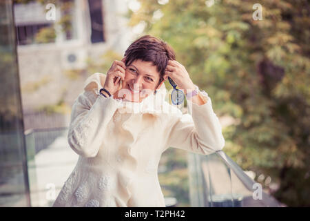 D'âge moyen à la mode mature woman wearing white robe chemisier ou laine noir lunettes de soleil est maintenant parler, parler sur un smartphone gaiement laughin Banque D'Images