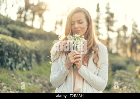 Femme heureuse avec de longs cheveux rouges et un agréable sourire dans les rayons du soleil est titulaire d'un bouquet de fleurs délicates en face d'elle et aime Banque D'Images