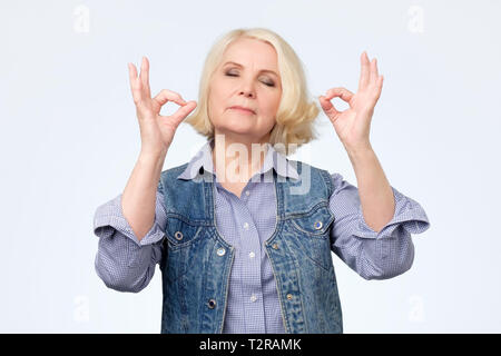 Senior woman smiling with eyes closed faisant le geste de la méditation avec les doigts. Concept de yoga. Banque D'Images