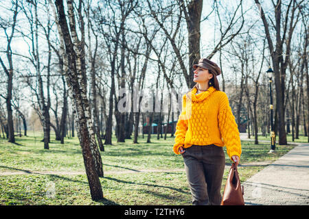 Jeune femme tenant un sac à main élégant et chandail jaune tout en marchant dans le parc. Femme printemps Vêtements et accessoires. Fashion Banque D'Images