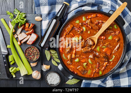 Boeuf et à la bière irlandaise traditionnelle ragoût avec carottes, céleri, carottes et des épices dans une cocotte avec des ingrédients et une bouteille de bière sur un w noir Banque D'Images