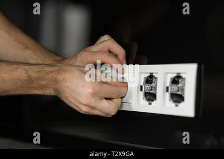 Close-up d'une main de l'électricien sans outils et gants de démontage d'un prise électrique blanc sur un mur de verre noir. Banque D'Images