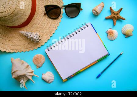 La mise en page sur le thème de vacances à la mer : chapeau de paille, lunettes de soleil, coquillages et bloc-notes avec stylo sur un fond bleu, vue du dessus Banque D'Images