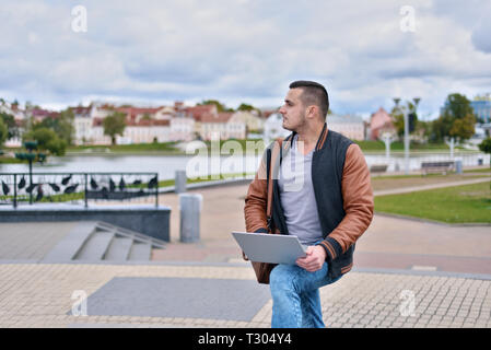 Les jeunes en blouson de cuir avec succès pigiste travaillant dans la rue. homme regarde pensivement sur le côté. le mec est tenue de l'ordinateur portable sur ses genoux Banque D'Images