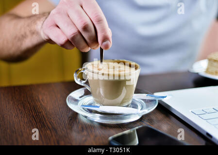 L'homme est en remuant cuillérée de sucre dans la tasse de cappuccino. sur la table est smartphone et ordinateur portable. châssis fermer Banque D'Images