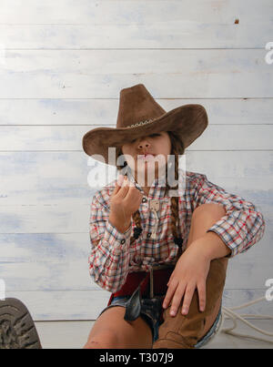 Une petite fille dans un chapeau de cow-boy à larges bords portant un costume traditionnel et de hautes bottes avec un lasso est en train de manger une paille sur un fond en bois clair. Banque D'Images