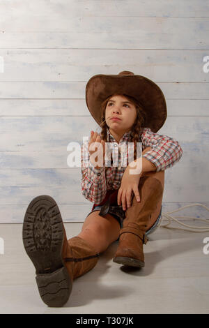 Une petite fille dans un chapeau de cow-boy à larges bords portant un costume traditionnel et de hautes bottes avec un lasso est en train de manger une paille sur un fond en bois clair. Banque D'Images