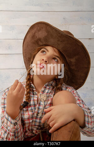 Une petite fille dans un chapeau de cow-boy à larges bords portant un costume traditionnel et de hautes bottes mange une paille sur un léger fond de bois Banque D'Images