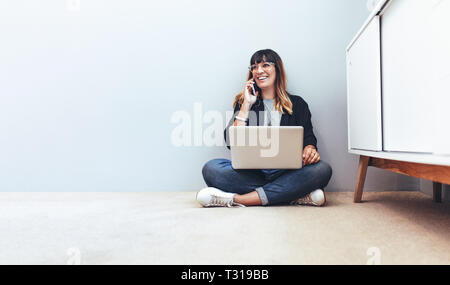 Femme entrepreneur gère ses affaires à la maison. Freelancer sitting on floor travaillant sur un ordinateur portable et de conversations au téléphone mobile. Banque D'Images