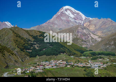 Kazbek de montagne avec de la neige blanche, ville de Stepantsminda pointe à l'avant-plan et l'église de trinité Gergeti à gauche avec le ciel bleu au-dessus. Banque D'Images