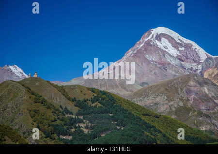 Kazbek montagne couverte de neige blanche avec PIC et l'église de trinité Gergeti en premier plan avec ciel bleu au-dessus. Banque D'Images