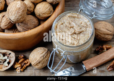 Pot en verre de beurre de noix biologiques crus ou de pâte, des écrous dans bol en bois et le couteau sur la table de cuisine. Vue d'en haut. Banque D'Images