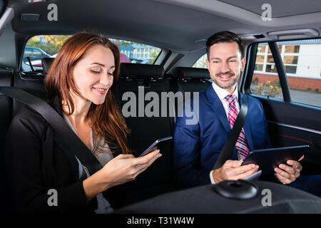 Happy Young Businesswoman Sitting à l'intérieur de voiture à côté de son collègue masculin Using Smartphone Banque D'Images