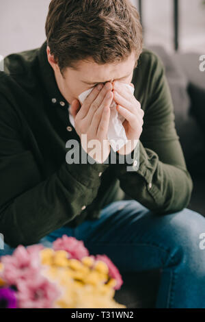 L'homme avec l'allergie au pollen couvrant visage pendant l'éternuement dans les tissus à proximité de fleurs Banque D'Images