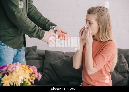 Portrait d'homme tenant un verre d'eau et de comprimés près de blonde femme avec l'allergie au pollen l'éternuement dans les tissus à proximité de fleurs Banque D'Images