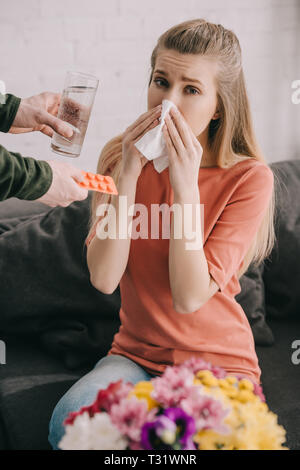 Portrait d'homme tenant un verre d'eau et de comprimés près de femme avec l'allergie au pollen l'éternuement dans les tissus à proximité de fleurs Banque D'Images