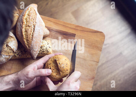 Gros plan Vue de dessus de l'homme de race blanche mains couper un morceau de pain avec un couteau de cuisine avec woodboard en arrière-plan la lumière naturelle Banque D'Images