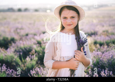 Jolie fille aux cheveux longs dans une robe de lin et un chapeau avec un bouquet de lavande debout dans un champ de lavande Banque D'Images