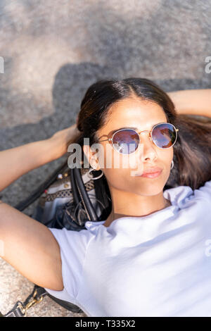 Portrait d'une belle jeune femme dans une chemise blanche et des lunettes en position allongée sur une surface en béton sur une journée ensoleillée. Banque D'Images