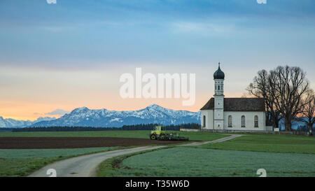 Matin l'agriculture en terrain alpin vert avec le tracteur et petite église sur fond de montagnes enneigées des Alpes Banque D'Images