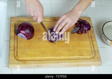 Vue aérienne de la mains commençant à couper la moitié des ​Red l'oignon sur une planche à découper, un bol d'eau sur le côté. Banque D'Images