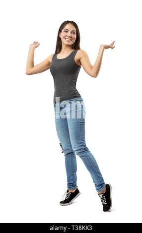 Young smiling woman in sleeveless top gris et bleu jeans debout sur la pointe des pieds avec les bras pliés et soulevé à côtés isolé sur fond blanc. Banque D'Images