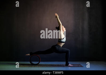 Jeune femme pratiquant le yoga, debout dans un exercice guerrier, Virabhadrasana je pose, à l'aide de roue, portant des vêtements noirs. Côté, pleine longueur Banque D'Images