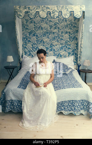 Attractive young woman in dress regency authentique assis sur un lit d'époque Banque D'Images