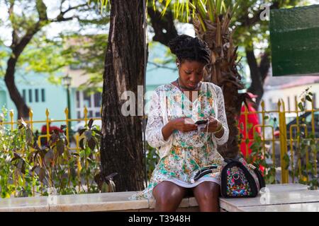 Belle Jeune femme créole avec la peau foncée assis dans jolie robe à fleurs à écouter de la musique et de la messagerie texte sur appareil mobile Banque D'Images