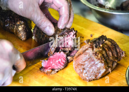 Chef de mains dans la préparation des aliments à usage unique, des gants en plastique de surlonge de boeuf rôti cuit découper avec un grand couteau sur une planche à découper en bois. Services de restauration Banque D'Images