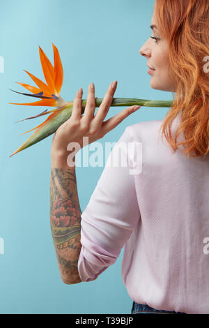 Fille avec un tatouage tenant sur son épaule une fleur orangée strelitzia autour d'un fond bleu avec l'espace pour le texte. Mise en page créative Banque D'Images